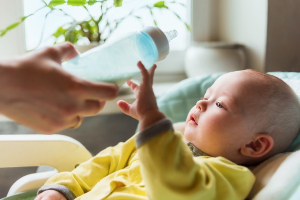 Ein Baby liegt in einer Wippe und greift nach einer Babyflasche mit Tee, die ihm hingehalten wird.
