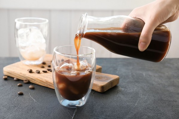 Ein Glas mit Eiswürfel wird mit kalt aufgebrühten Kaffee aus eine Flasche aufgefüllt. Im Hintergrund liegt ein Holzbrett auf dem ein leeres Glas mit Eiswürfeln befindet.