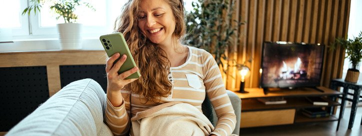 Eine Schwangere sitzt auf dem Sofa und schaut lächelnd auf ihr Handy.