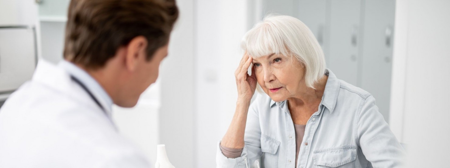 Eine Seniorin sitzt im Behandlungszimmer einer Arztpraxis. Ihr gegenüber sitzt ein Arzt im Gespräch der mitschreibt. Die Seniorin fässt sich während des Gesprächs mit der rechten Hand an die Schläfe.