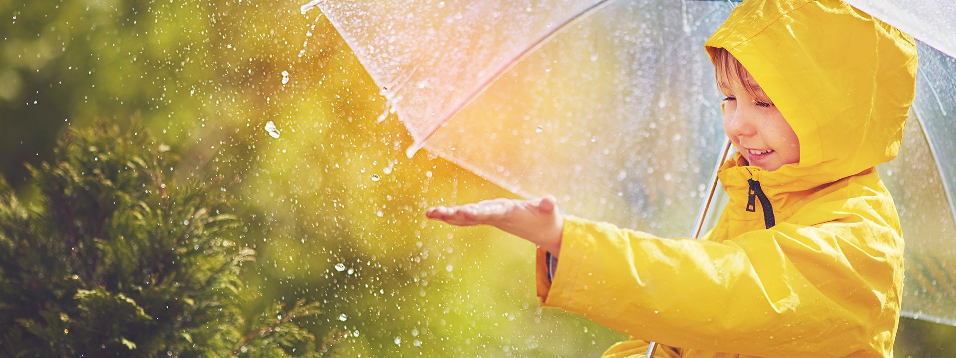 Ein circa 5-jähriger Junge mit gelben Regenmantel und Regenschirm genießt den Regen.