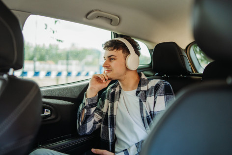 Ein etwa 20-jähriger Mann sitzt hinten im Auto und schaut verträumt aus dem Fenster. Dabei trägt er Kopfhörer.