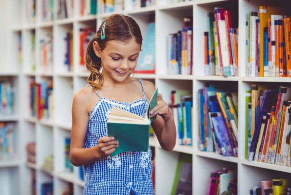 Ein etwa 8-jähriges Mädchen steht in der Bibliothek vor einem Bücherregal und blättert lächelnd in einem Buch.