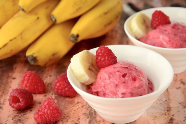 Auf einem Holztisch steht eine Schale Himbeer-Nicecram. Außerdem liegen frische Bananen und Himbeeren auf dem Tisch.