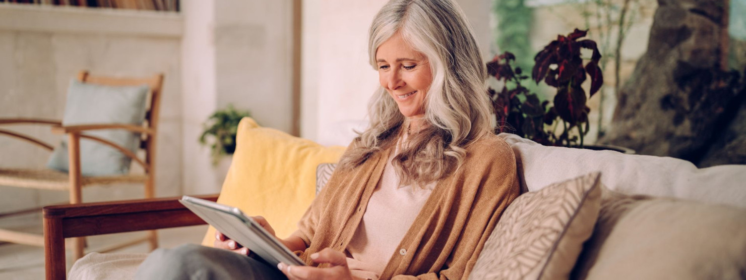 Eine Seniorin sitzt auf ihrem Sofa und schaut glücklich auf ihre Tablet.