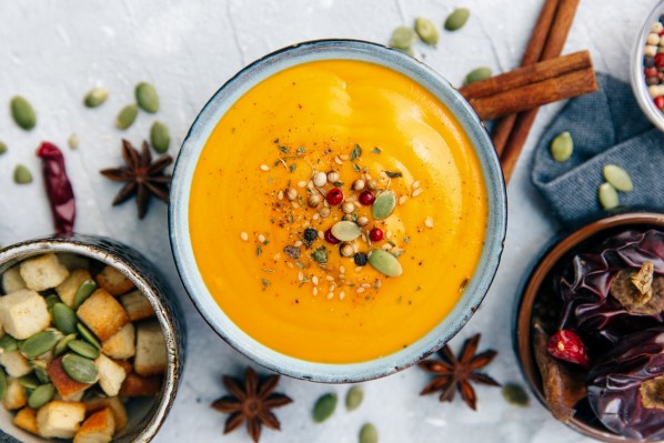 Blick von oben auf eine Schüssel mit Kürbissuppe. Sie ist garniert mit Kräutern, Gewürzen, Kürbiskernen und Beeren. Um die Suppenschüssel stehen Schüsseln mit Croutons und Kürbiskernen, bunten Pfefferkörnern und Beeren. Auf dem Tisch liegen zwei Zimtstangen, Anissterne und Kürbiskerne verstreut.