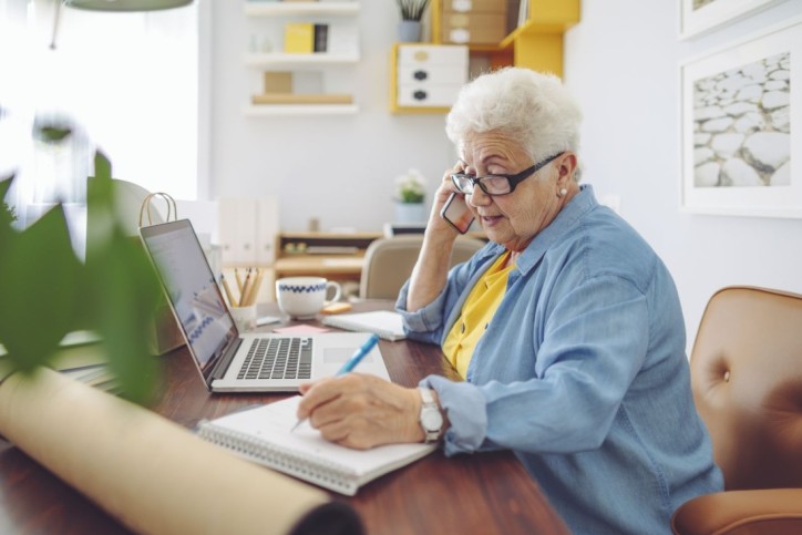 Eine Seniorin sitzt am Laptop, telefoniert und schreibt parallel etwas auf.