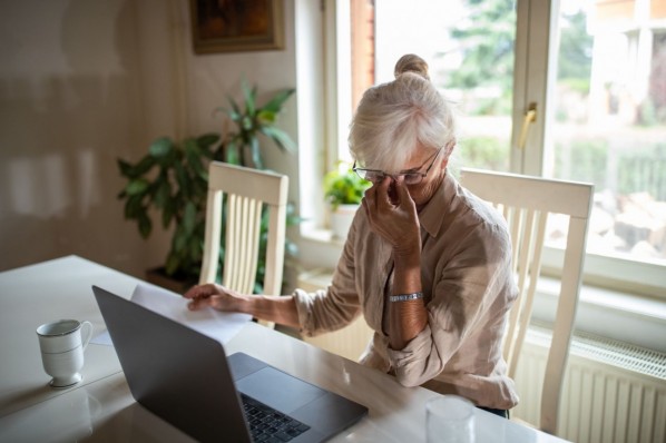 Eine Seniorin sitzt an einem Schreibtisch und reibt sich die Augen. Vor ihr steht ein Laptop.