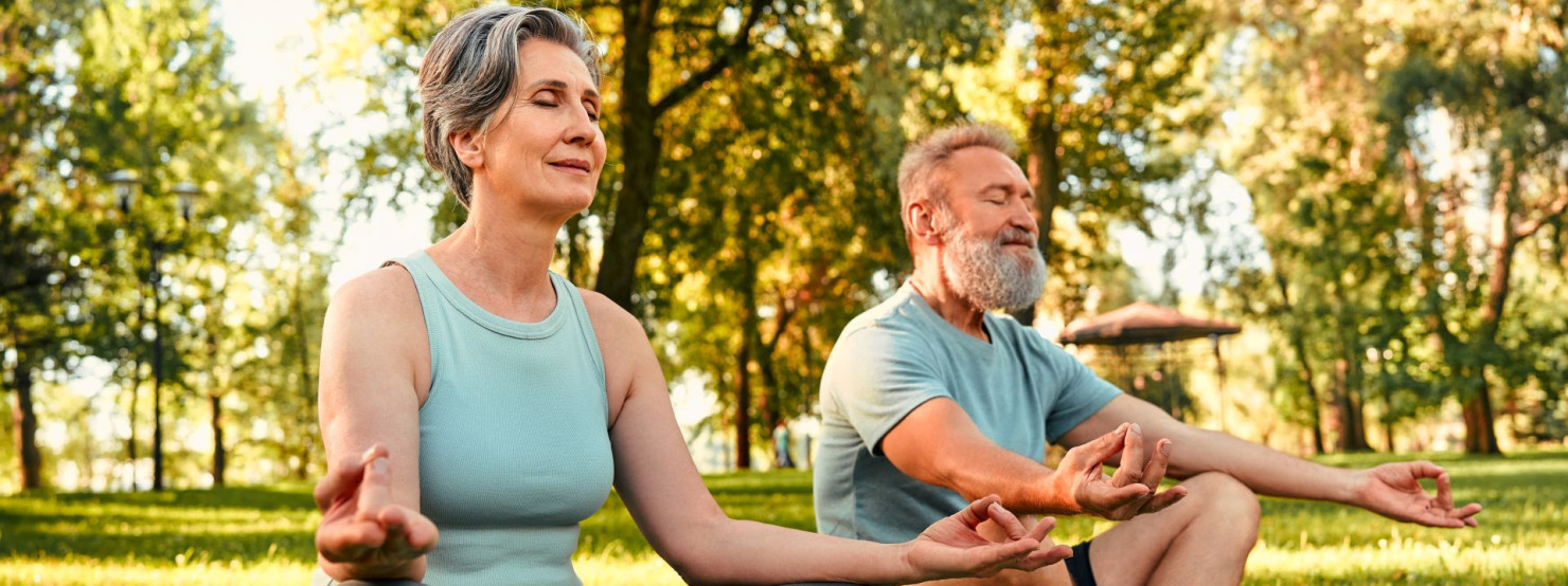 Ein Paar sitzt in einem Park auf ihren Yogamatten und machen Yoga mit der AOK Yogaschule.