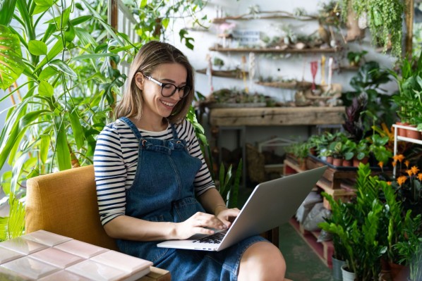 Eine etwa 30 jährige Frau sitzt einem Raum umgeben von Pflanzen. Auf dem Schoß hat sie einen Laptop auf dem sie arbeitet. 