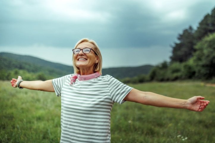 Eine Frau steht mit ausgebreiteten Armen in der Natur und lächelt.
