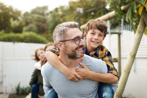Ein Vater trägt seinen etwa 6 jährigen Sohn auf dem Rücken. Beide schauen sich an und lächeln.