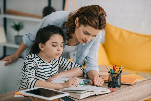 Eine Mutter hilft ihrer etwa 8 jährigen Tochter bei Hausaufgaben. Dabei beugt sie sich von über ihre Tochter und schaut auf ein Buch.