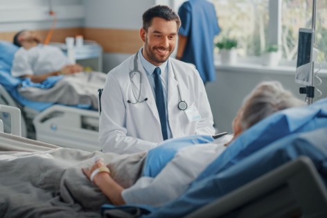 Ein etwa 40-jähriger Arzt sitzt lächelnd am Bett eines Krankenhauspatienten.