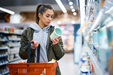 Eine etwa 25-jährige Frau steht im Supermarkt vor dem Produktregal und liest die Rückseite eines Produktes.