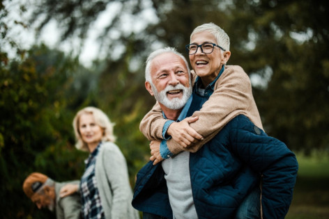Älteres Paar, das Spaß beim Huckepack im Park hat. Ihre Freunde sind im Hintergrund