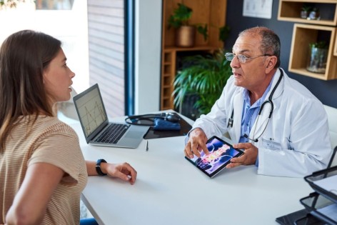 Eine Patientin und ein Orthopäde besprechen sich in einem Arztzimmer. Der Orthopäde zeigt der Patientin etwas auf seinem Tablet.