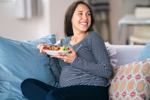 Eine etwa 30-jährige Frau lächelt beim Essen auf der Couch.