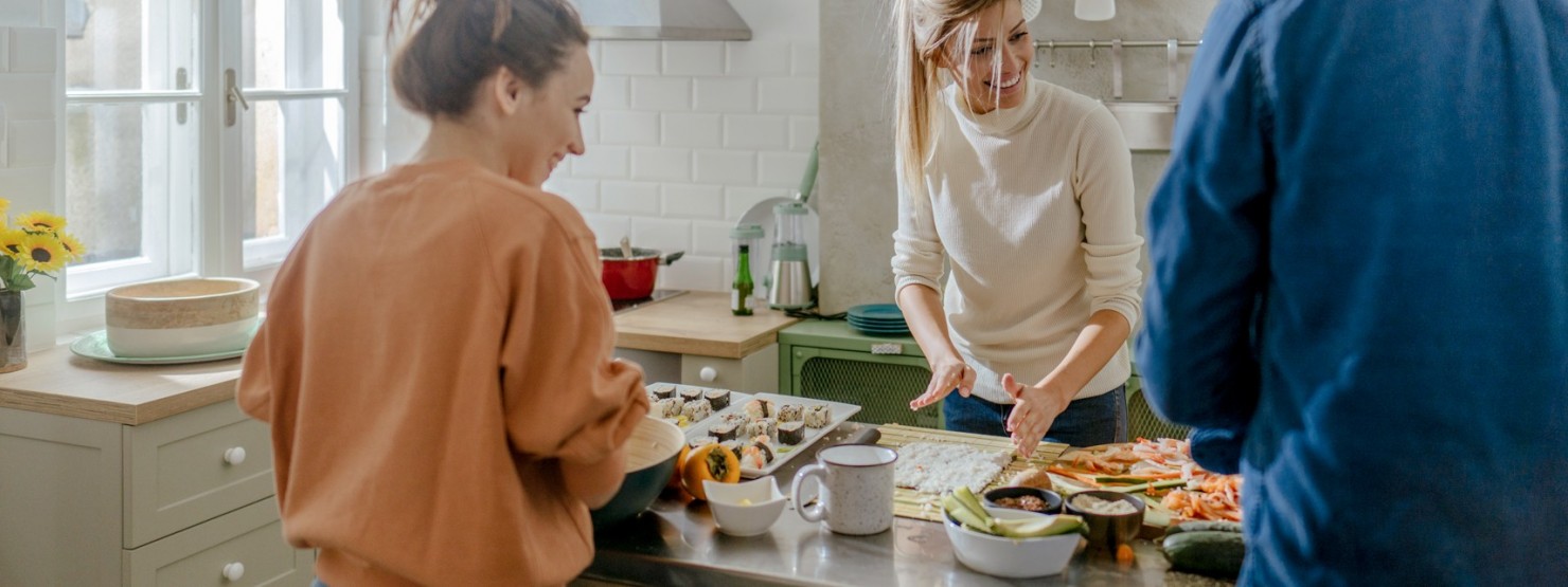 Zwei Frauen, beide etwa 20 Jahre alt, und ein Freund machen zusammen in der Küche Sushi.