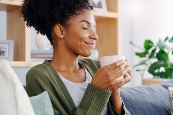 Eine etwa 30-jährige Frau sitzt lächelnd mit geschlossenen Augen und einer Tasse auf dem Sofa.