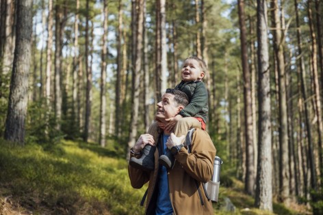 Ein Vater trägt seinen etwa 2 jährigen Sohn auf den Schultern durch den Wald.
