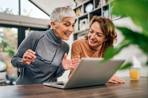 Eine Seniorin mit einer Brille in der Hand und eine etwa 40 jährige Frau sitzen an einem Tisch. Vor ihnen steht ein Laptop und beide lächeln. 