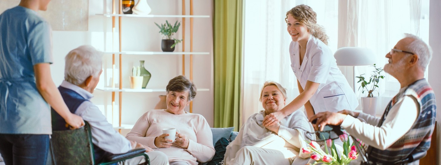 Vier Senioren und Seniorinnen sitzen zusammen. Neben ihnen stehen zwei Pflegerinnen. 