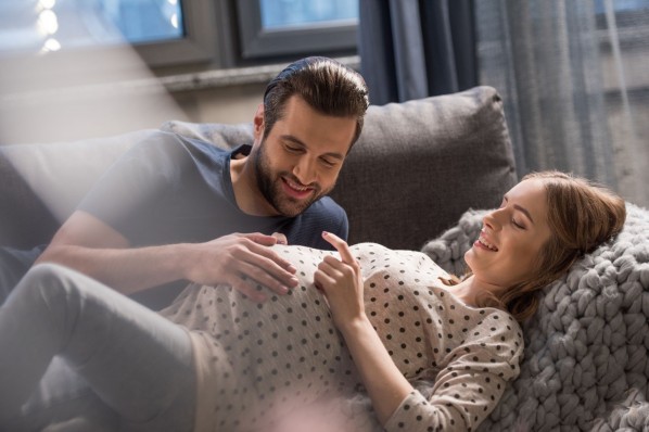 Ein Pärchen liegt gemeinsam auf dem Sofa. Die Frau ist schwanger und beide lächeln. Der Mann berührt den Babybauch der Frau.