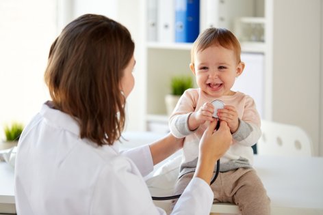 Ein etwa einjähriges Kind wird von einer Kinderärztin mit dem Stethoskop untersucht. Das Kleinkind lacht ausgiebig.
