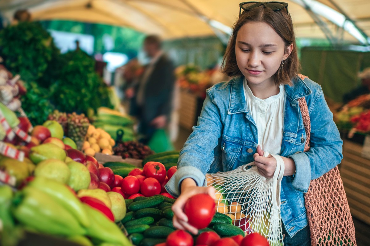 Etwa 20-jährige Frau legt auf einem Markt eine Tomate in ihr Gemüsenetz.