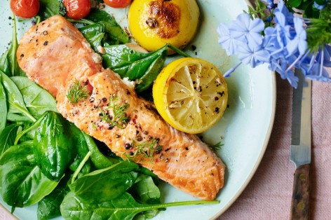 Gegrillter Lachs liegt zusammen mit Zitronen, gegrillten Tomaten und frischen Blattspinat auf einem hellen Teller.