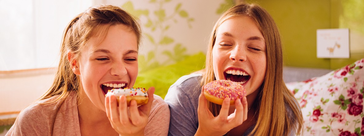 Zwei ca 12-jährige Mädchen sitzen im Kinderzimmer auf dem Bett und essen einen Donut