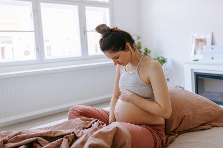 Eine schwangere Frau hält ihren Bauch und sitzt auf dem Bett.