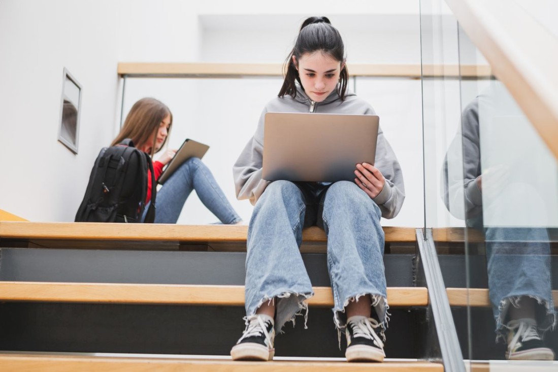 Zwei weibliche Jugendliche sitzen im Schultreppenhaus und schauen auf ihren Laptop.