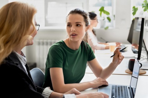 Eine etwa 30 jährige Frau unterhält sich an ihrem Schreibtisch im Büro mit ihrer Kollegin. Sie haben einen Laptop vor sich stehen.