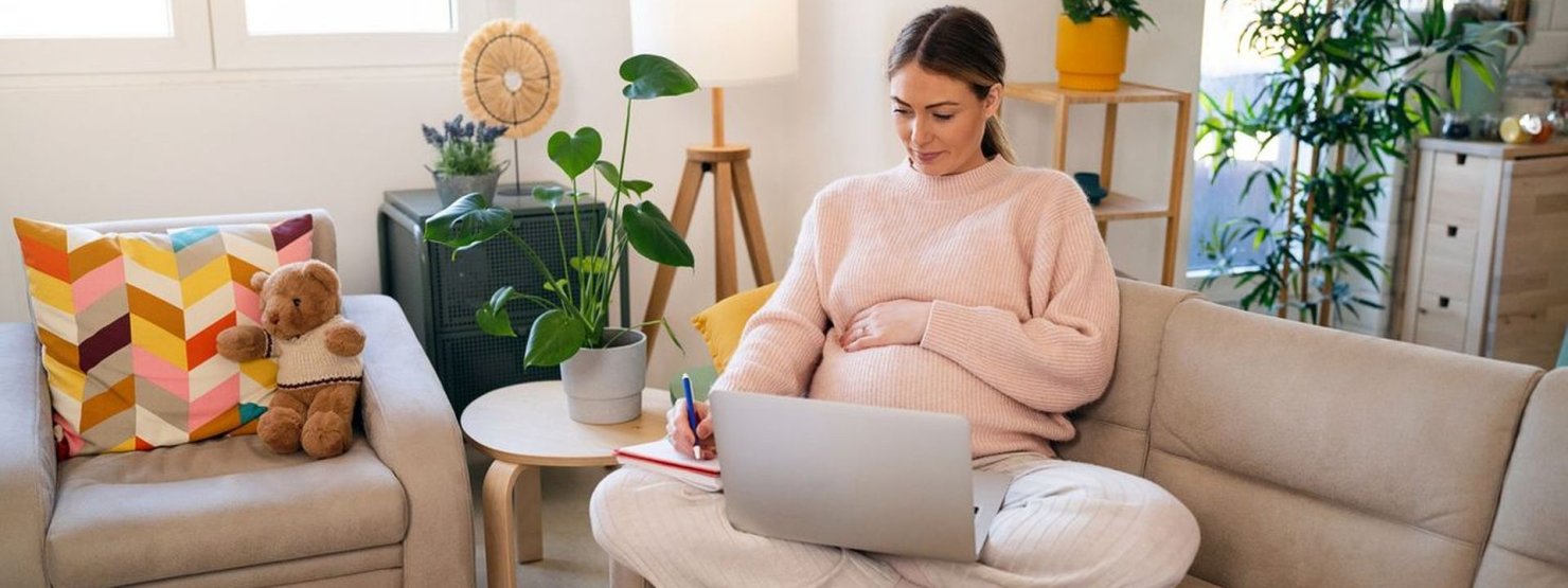 Eine circa 25-jährige, schwangere Frau sitzt mit Laptop auf der Couch.