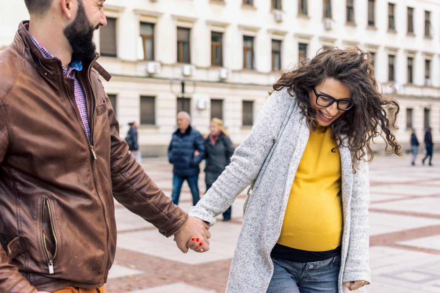 Ein junges Paar spaziert Hand in Hand über einen Platz in der Stadt. Sie ist schwanger.