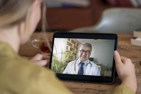 Eine Frau hält ein Tablet in der Hand. Auf dem Bildschirm ist ein Arzt zu sehen, der die Frau berät.