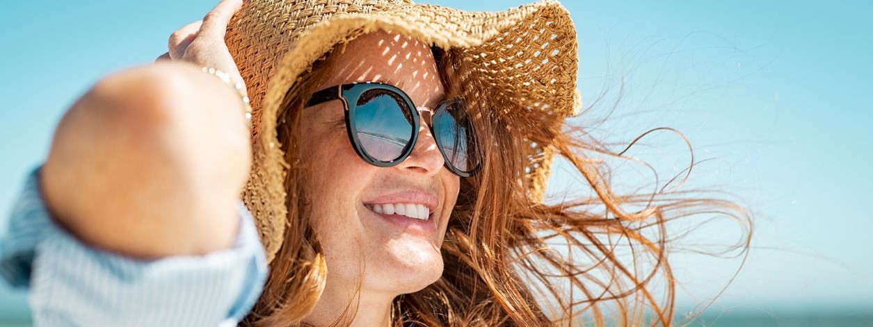 Eine etwa 40 jährige Frau mit Sonnenbrille und Sonnenhut sonnt sich am Strand. Dabei lächelt sie und schaut in die Ferne.