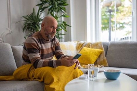 Ein etwa 55-jähriger Mann sitzt auf dem Sofa und schaut auf sein Handy.