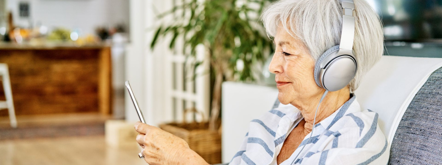 Eine Seniorin hört über ihre Kopfhörer den AOK Podcast Pflege. Dabei schaut sie auf ihr Smartphone und lächelt.