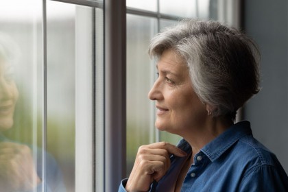 Ältere Frau Anfang 60 schaut mit einem leichten Lächeln aus dem Fenster