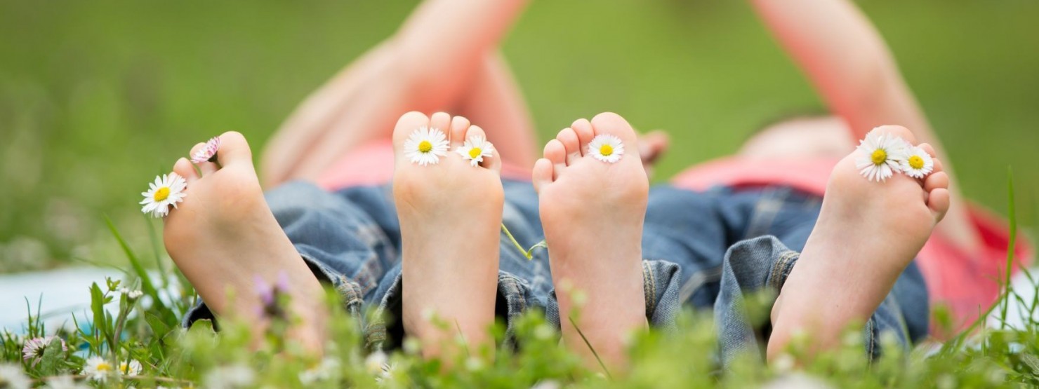 Zwei Kinder liegen auf einer Wiese. Sie sind barfuß, zwischen ihren Zehen stecken Gänseblümchen.