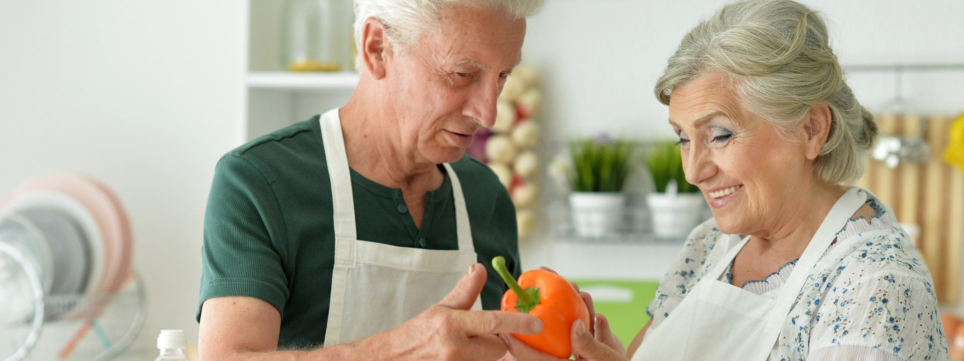 Ein Seniorenpaar steht gemeinsam in der Küche und kocht. Die Frau hält eine Paprika in der Hand. Beide lächeln.