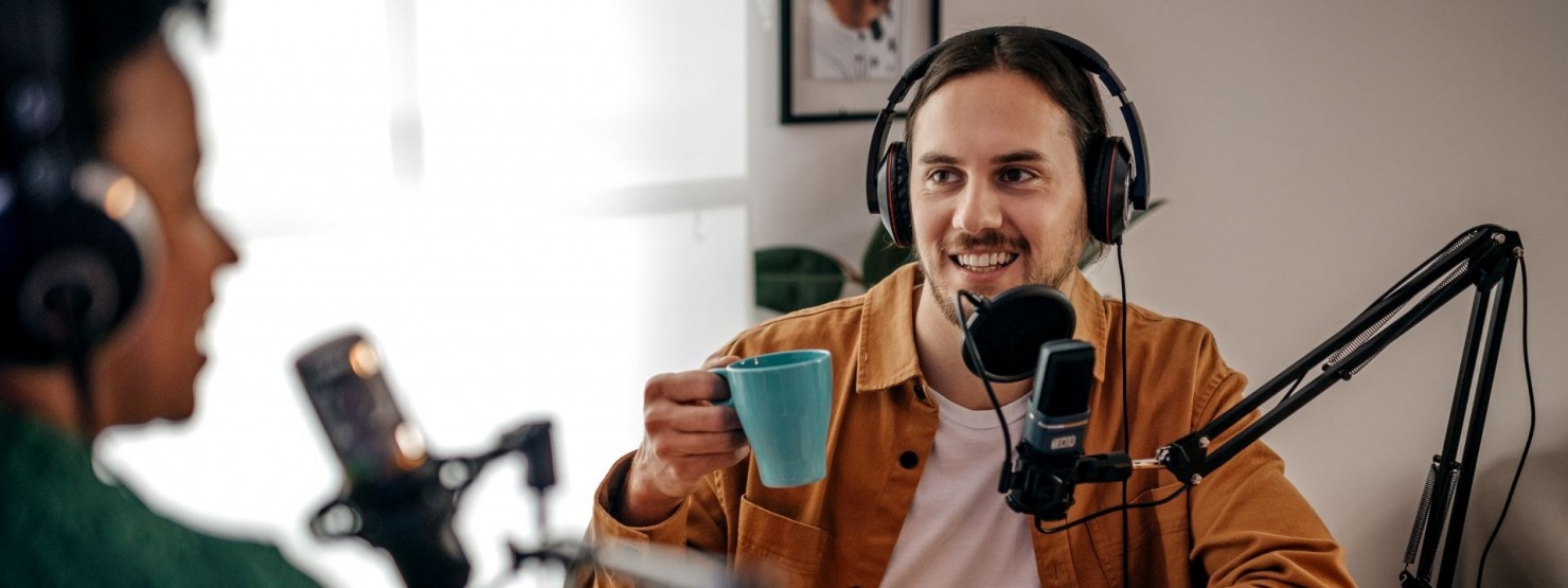 Zwei Männer sitzen sich an einem Tisch gegenüber und nehmen über Mikrofone eine Podcastfolge auf.