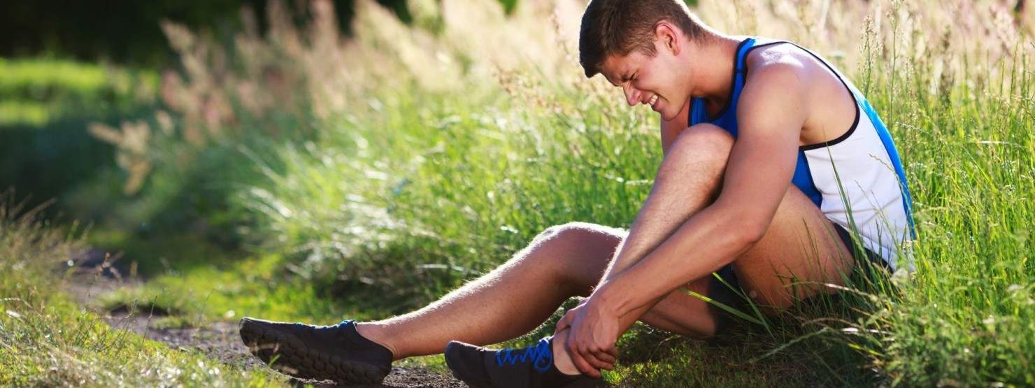 Ein etwa 25 jähriger Sportler sitzt am Rand eines Wiesenwegs und hält sich den linken Knöchel. Dabei verzieht er sein Gesicht vor Schmerzen.