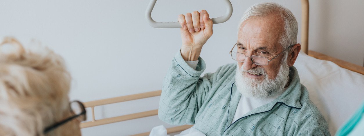 Ein Senior liegt in einem Pflegebett zuhause und spricht mit seiner Ehefrau. Ein Pfleger stellt das Pflegebett mit einer Fernbedienung ein.