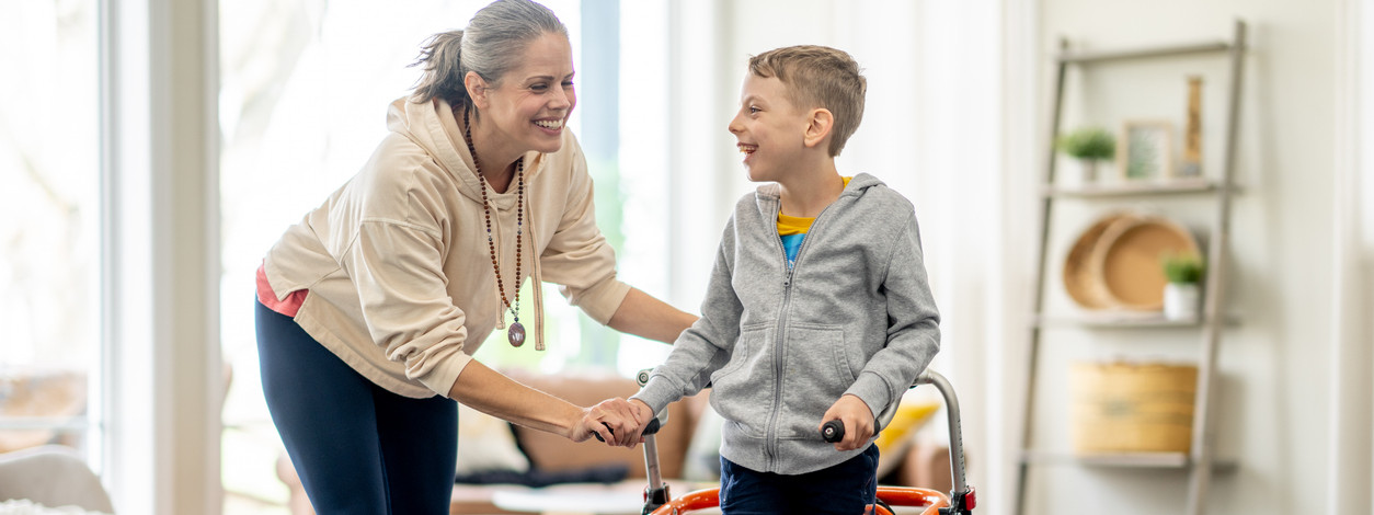 Ein Junge mit Gehbehinderung steht in einer medizinischen Stehhilfe. Seine Mutter unterstützt ihn zusätzlich.