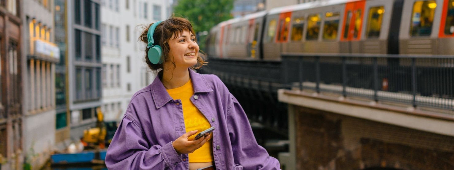 Eine etwa 20-jährige Frau hört einen Podcast über ihre Kopfhörer. Dabei steht sie mitten in der Stadt und wartet auf den Zug.