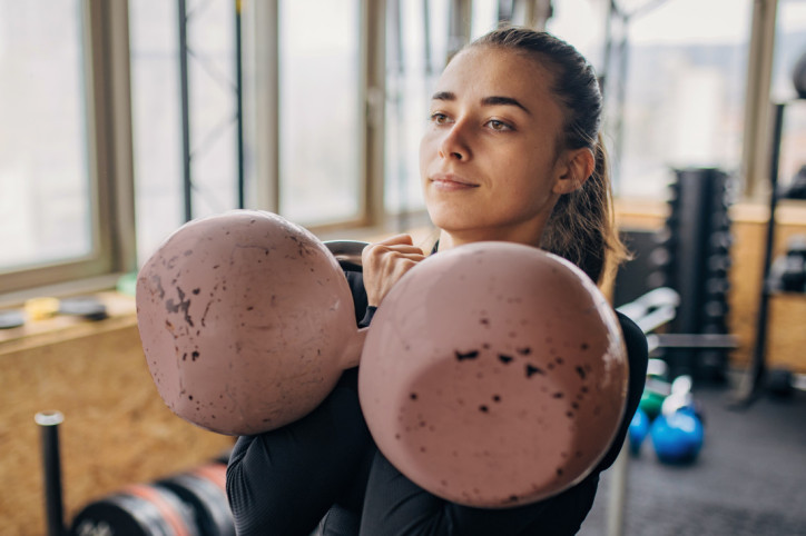 Eine etwa 18-jährige Frau hält zwei große Kugelhanteln in den Händen und blickt gelassen nach vorn. Sie befindet sich im Fitnessstudio.